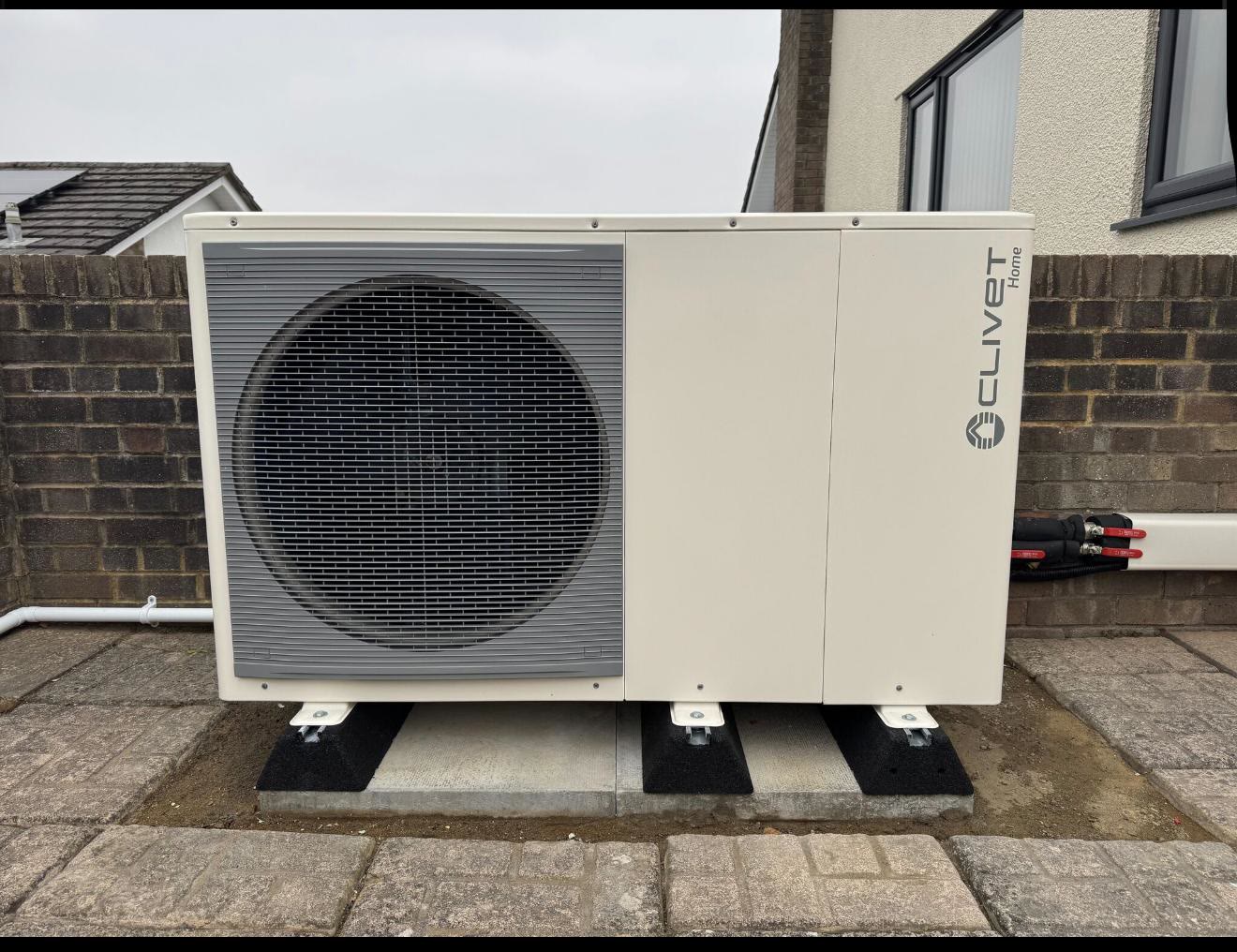 Clivet Home monobloc heat pump level on a poured concrete plinth beside a brick boundary wall, with insulated flexible connections